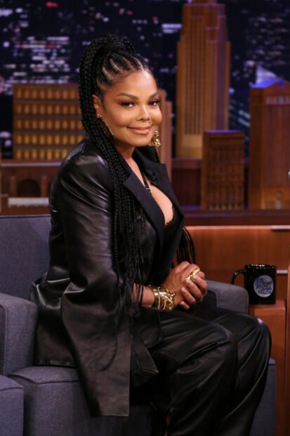 Janet Jackson wearing a black leather outfit with braided hair sitting on a talk show set