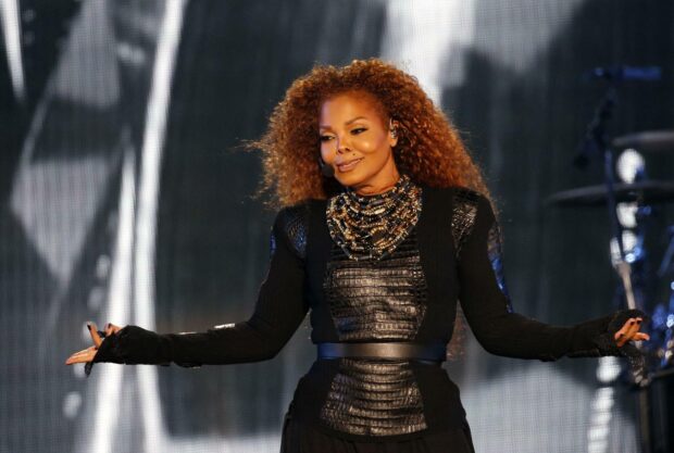 Janet Jackson wearing a black textured outfit with elaborate beaded necklace on stage
