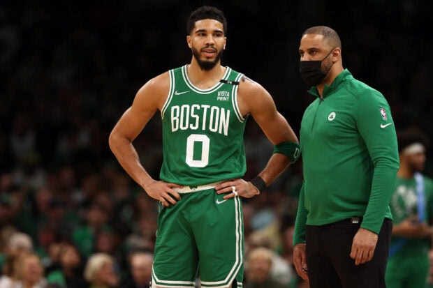 Jayson Tatum standing on the basketball court wearing a green Boston jersey talking to a coach
