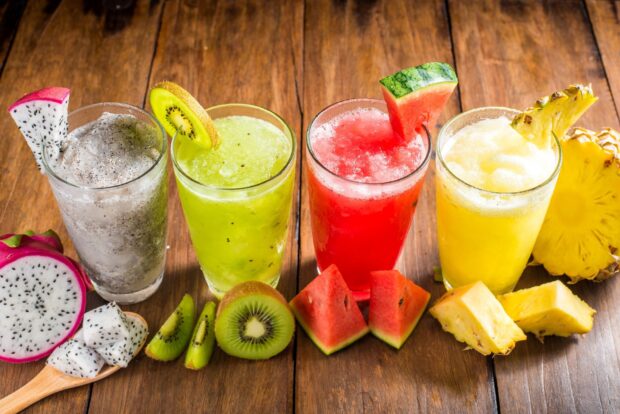 Four glass tumblers of kiwi dragon fruit watermelon and pineapple juices on a wooden table with fresh sliced fruits around