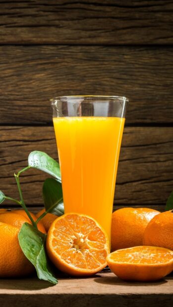 A glass of fresh orange juice surrounded by ripe oranges and green leaves on wooden surface