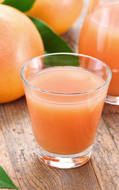 A glass of fresh grapefruit juice resting on a wooden table with ripe grapefruit fruits and green leaves visible