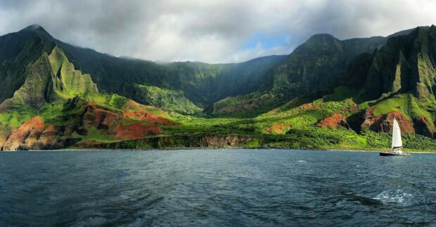 A lush green Kauai coastline with rugged cliffs and a sailboat on the ocean under cloudy skies