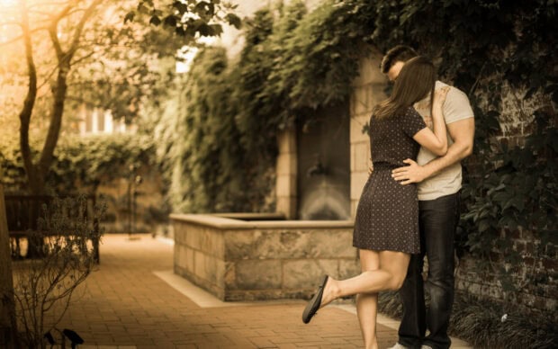 A couple sharing a kiss in an outdoor garden setting with the woman wearing a dark dress