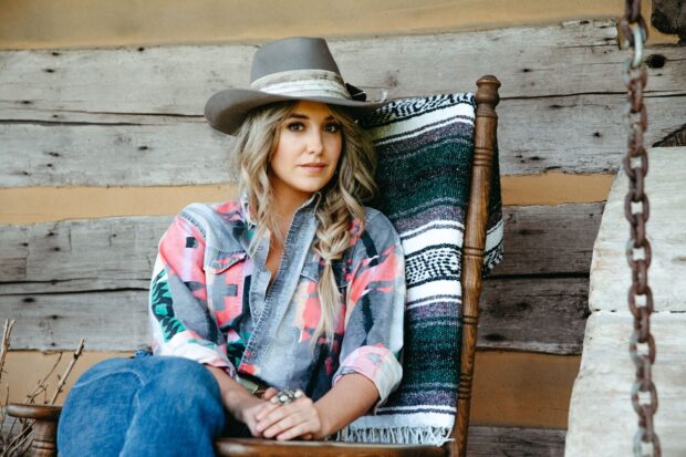 Lainey Wilson sitting on a wooden chair with a hat and colorful shirt in a rustic setting