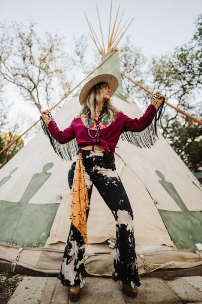 Lainey Wilson wearing a colorful western outfit with fringe standing in front of a teepee