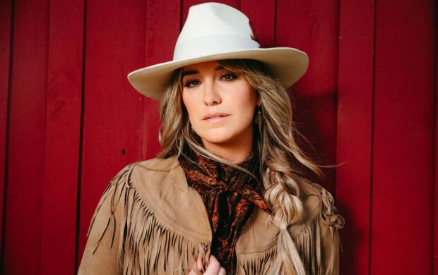 Lainey Wilson wearing a beige hat and fringe jacket standing against a red wooden wall