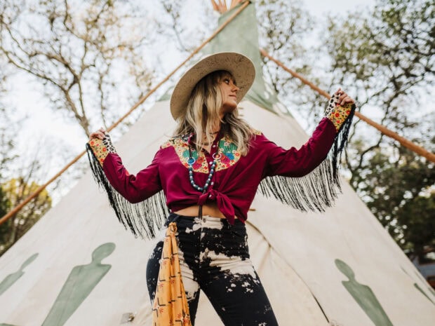 Lainey Wilson wearing a colorful western outfit and hat standing in front of a teepee