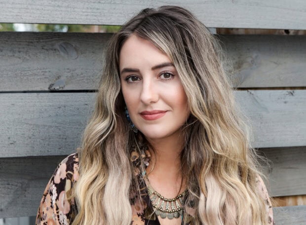 Lainey Wilson wearing a floral blouse with long blonde hair standing against a wooden fence