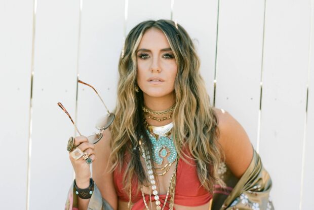 Lainey Wilson wearing bohemian jewelry and holding sunglasses in front of a white fence