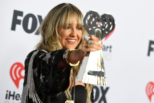 Lainey Wilson holding an award in a black outfit at an event background