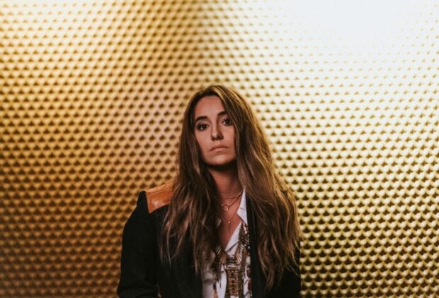 Lainey Wilson posing in front of a textured golden wall with long hair and layered necklaces