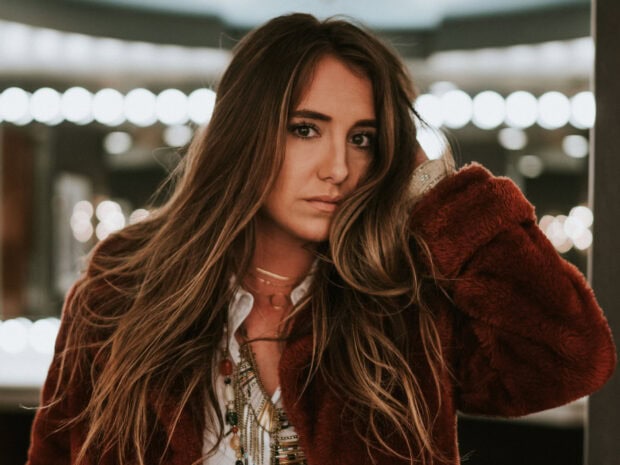 Lainey Wilson posing with long hair and layered necklaces in a cozy brown coat