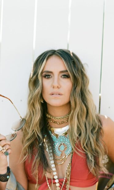 Lainey Wilson wearing layered necklaces and holding sunglasses in a close up portrait