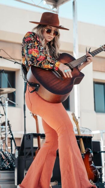 A woman wearing a hat and floral shirt playing guitar on stage captured for Lainey Wilson