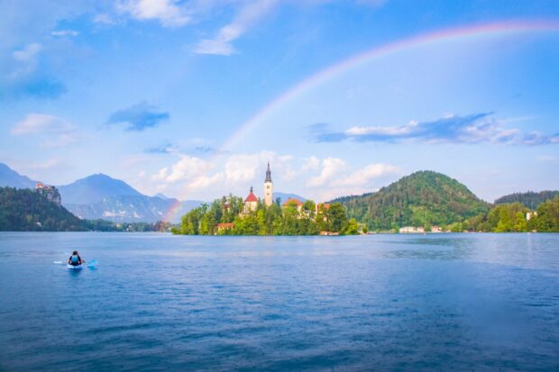 A scenic view of Lake Bled with a rainbow over the island and a person kayaking on the calm water