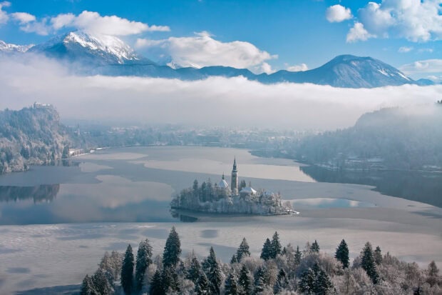 A snowy island and church surrounded by Lake Bled during winter with mountains in the background