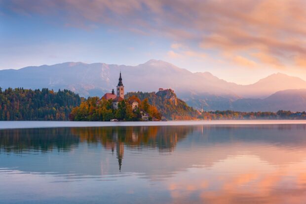 Lake Bled island with church and autumn trees reflecting on calm lake water at sunrise