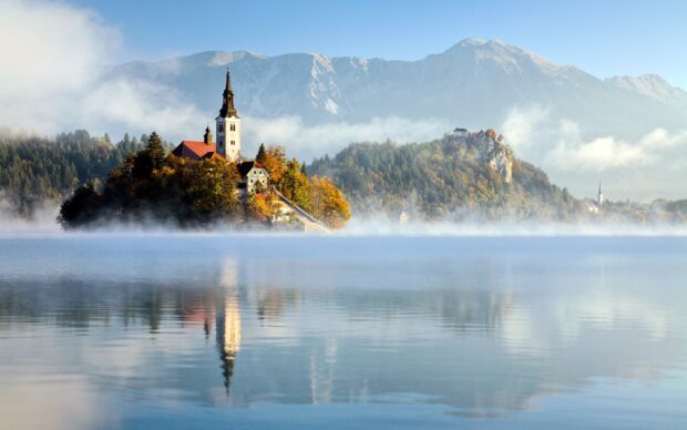 Lake Bled island with church and autumn trees surrounded by calm water and mountains in the background