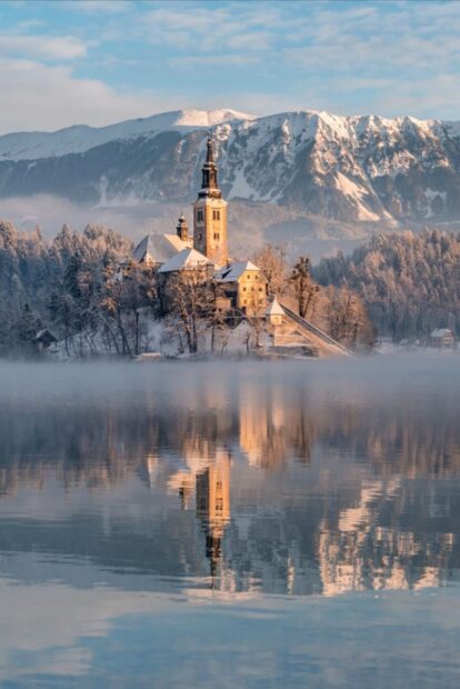 Winter scene of Lake Bled with snow covered church and mountains reflecting on the lake water