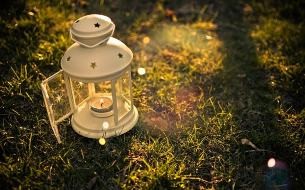 A white lantern with a lit candle inside placed on the grass in warm sunlight