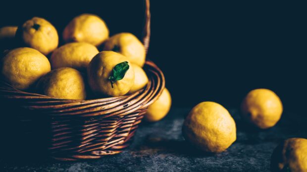 A basket full of fresh lemon fruit with one lemon showing a green leaf on a dark surface