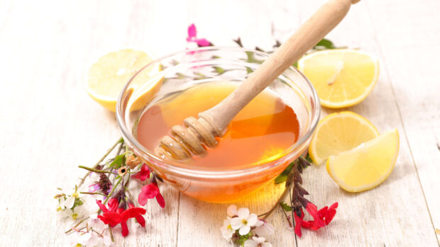 A clear bowl filled with honey surrounded by lemon slices and colorful flowers on a wooden surface