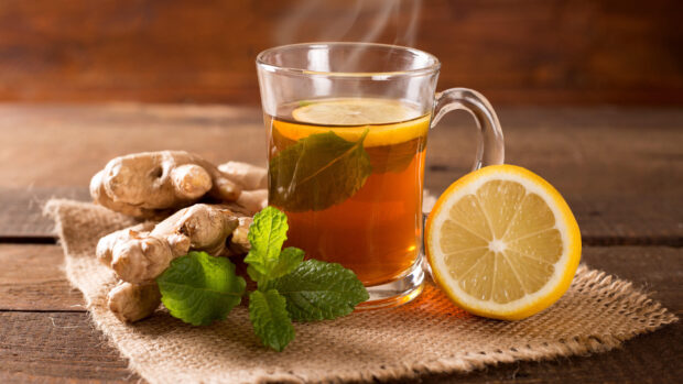 A lemon slice with fresh mint leaves next to ginger roots and a steaming cup of tea on a rustic table
