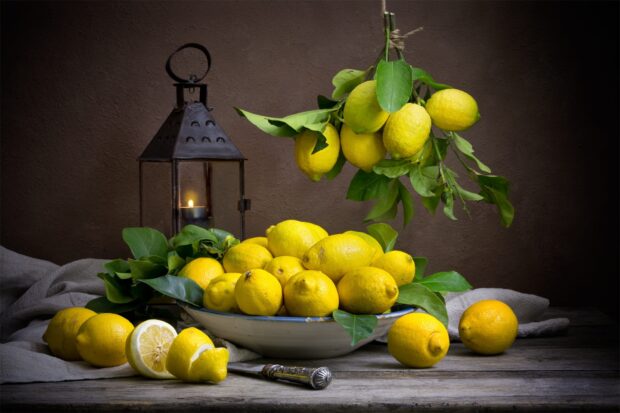 A bunch of lemons with green leaves arranged on a plate and hanging beside a lit lantern on wooden table