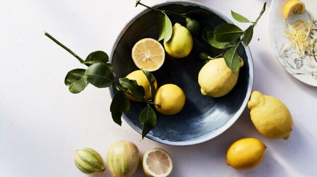 A bowl of fresh lemon with leaves and sliced citrus on a white surface
