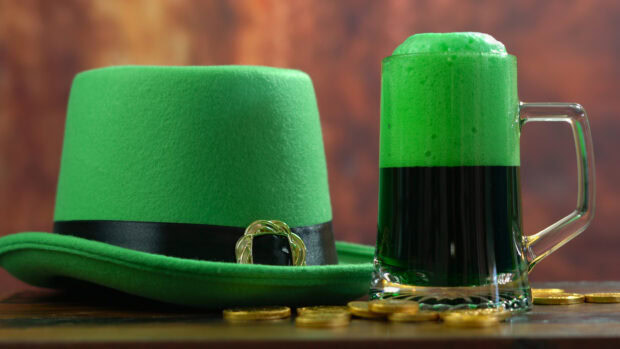 A green leprechaun hat with a black band and gold buckle next to a frothy green beer mug and scattered gold coins