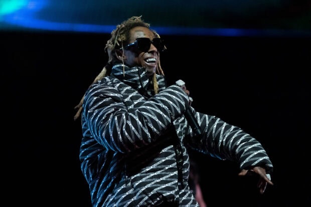 Lil Wayne wearing a patterned jacket and sunglasses performing on stage with a microphone in hand