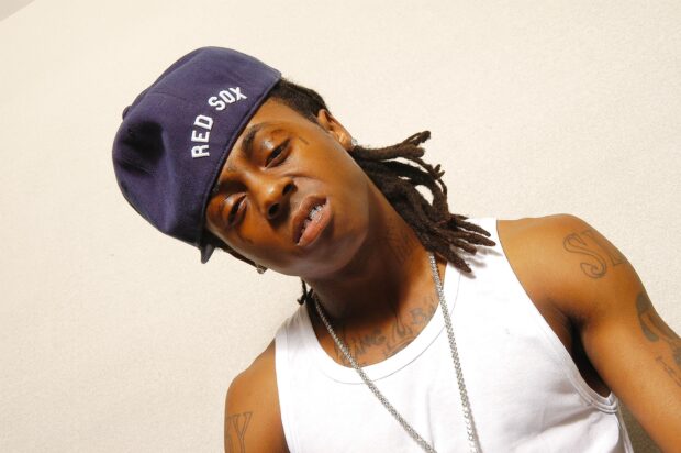 Lil Wayne with dreadlocks wearing a Red Sox cap and white tank top looking at the camera