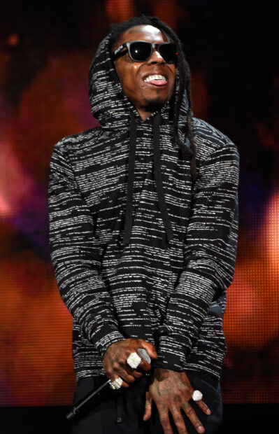 Lil Wayne wearing a patterned hoodie and sunglasses performing on stage