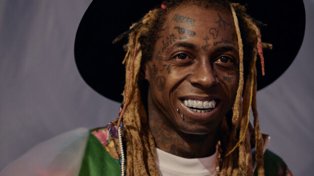 Lil Wayne with dreadlocks wearing a black hat and colorful jacket smiling
