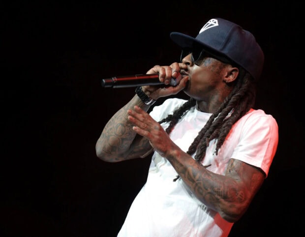 Lil Wayne performing on stage with detailed tattoos and dreadlocks wearing a cap and sunglasses