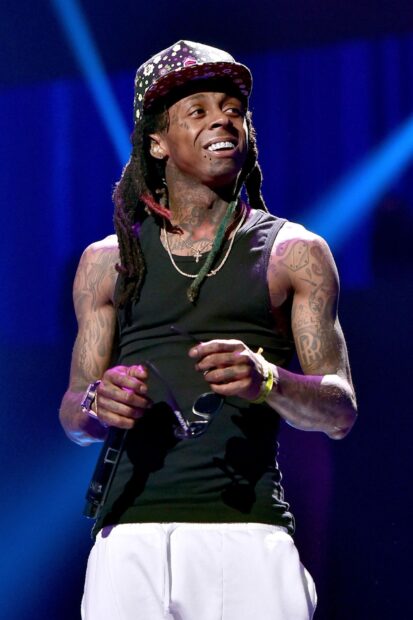 Lil Wayne wearing a colorful cap and black tank top holding sunglasses on stage