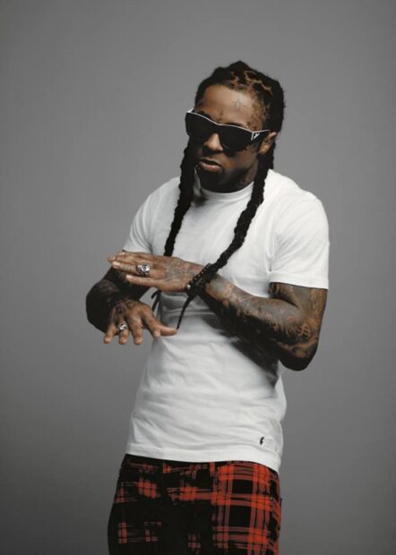 Lil Wayne posing with braided hair and sunglasses in a white shirt and red plaid pants