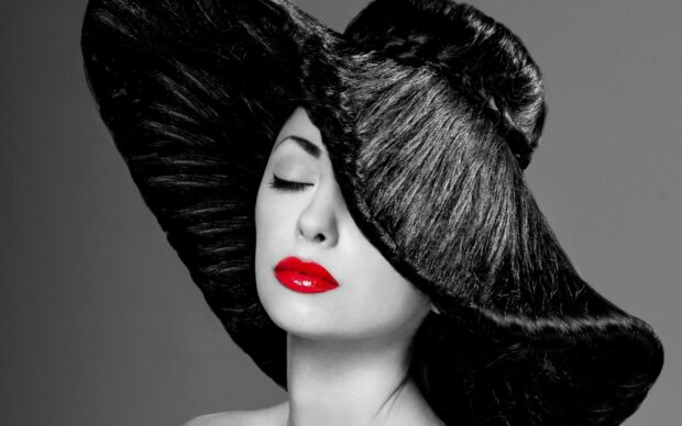 Woman with red lips wearing a large black hat and closed eyes using lipstick
