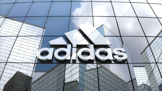 White Adidas logo displayed on glass building reflecting cityscape and sky clouds