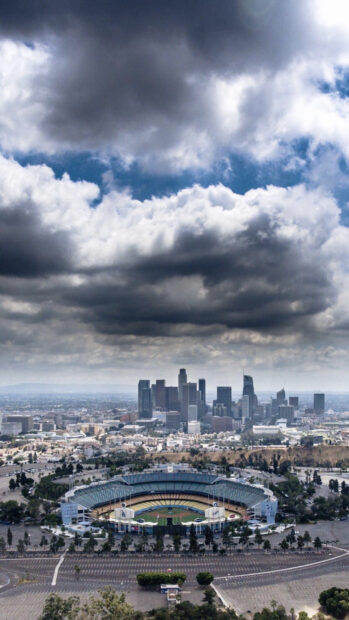 A panoramic view of Los Angeles skyline with a large baseball stadium in the foreground under cloudy skies