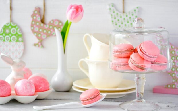Pink macaron with a spoon and more macarons displayed under a glass dome on a white wooden table