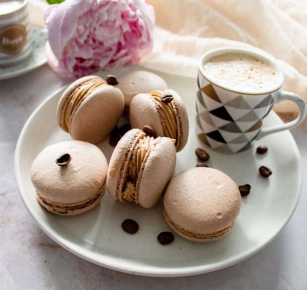 A plate of coffee flavored macaron with coffee beans and a cup of coffee on a light table