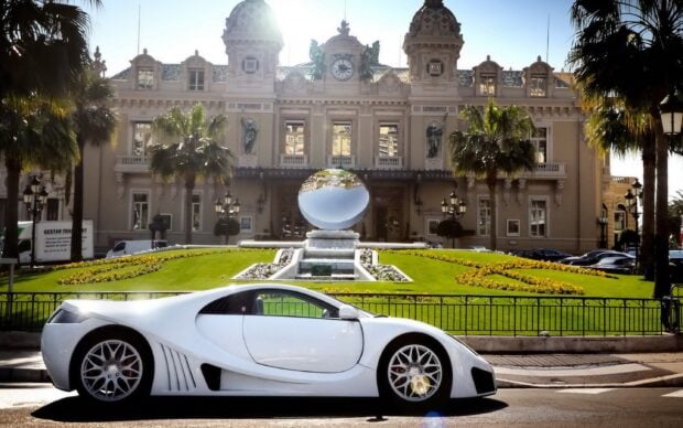 A luxury mansion with decorative sculptures and palm trees behind a white sports car on the street
