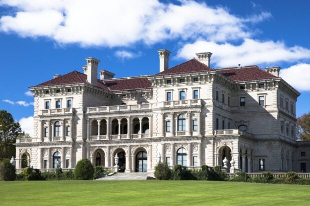 A grand mansion with intricate architectural details and a red tiled roof surrounded by green lawn and blue sky