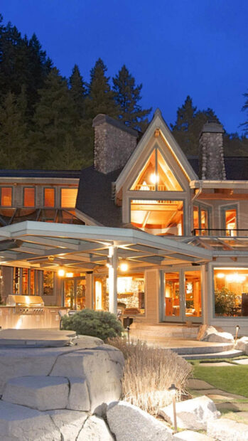A cozy mansion with stone chimneys and warm lights surrounded by trees and garden at night