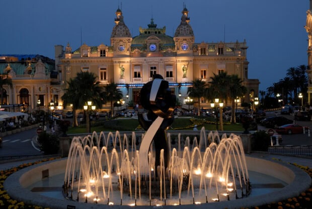 An illuminated fountain in front of a historic building in Monaco at dusk