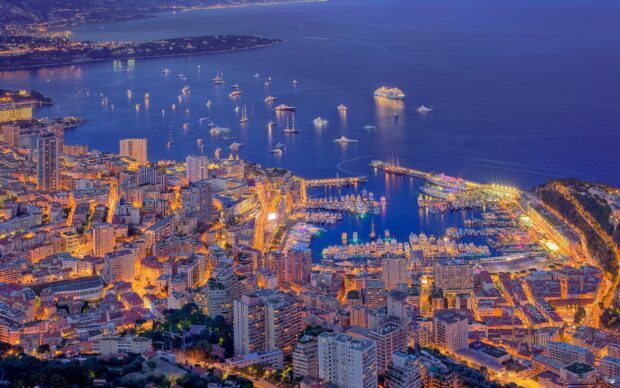 A panoramic view of Monaco cityscape at night with illuminated buildings and yachts in the harbor