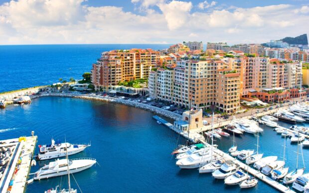 A scenic view of Monaco showing high rise buildings and luxury yachts in the harbor with clear blue sea