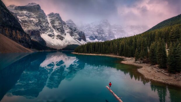 Snow covered mountains and forest reflected in Moraine Lake with a person standing on a log